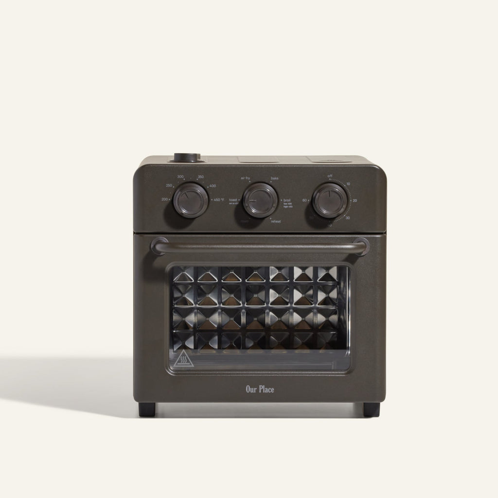 The AU - Wonder Oven is a dark gray countertop toaster oven with three top control dials and a front glass door featuring a diamond-patterned interior. "Our Place" appears at the bottom of the door. Doubles as an air fryer for extra versatility.