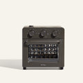 The AU - Wonder Oven is a dark gray countertop toaster oven with three top control dials and a front glass door featuring a diamond-patterned interior. "Our Place" appears at the bottom of the door. Doubles as an air fryer for extra versatility.